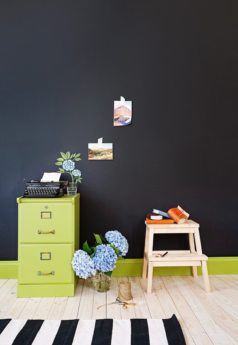 Hortensienstrauss zwischen grün lackiertem Büroschrank und Tritthocker vor schwarzer Wand
