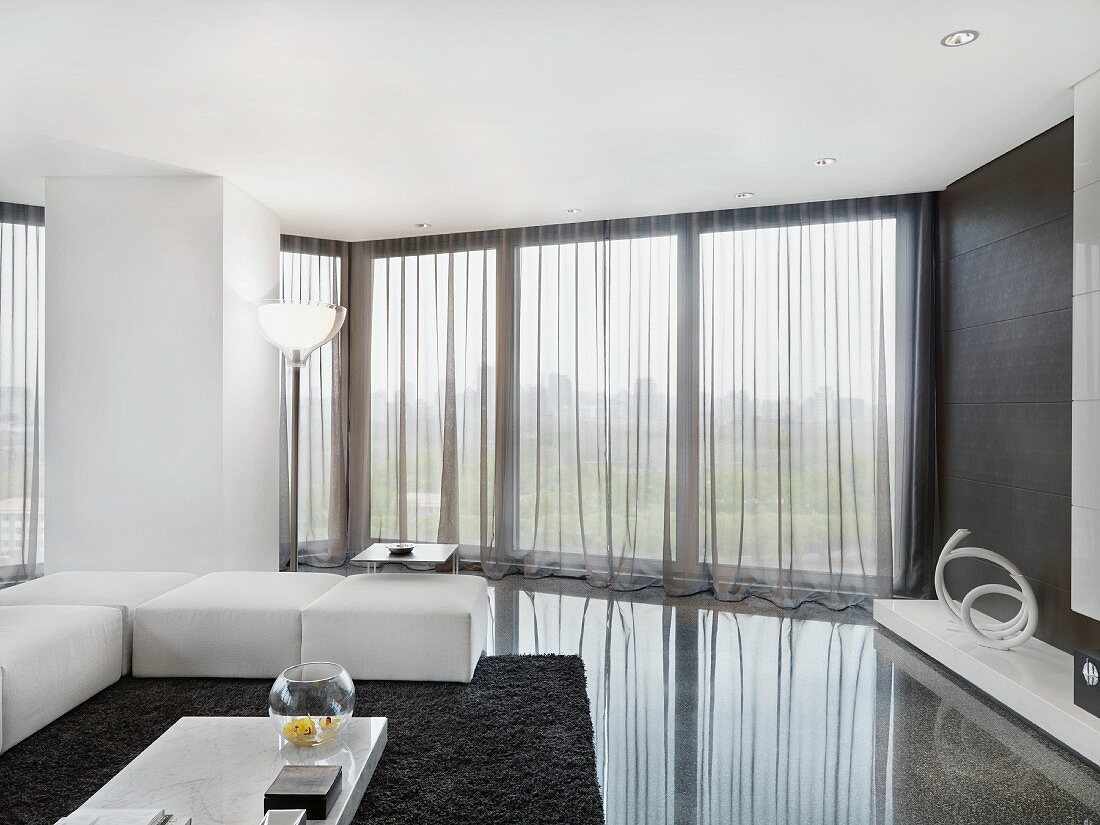 Elegant white upholstered living room furniture on a polished stone floor in a minimalist living room with see-through curtains in front of a bank of windows