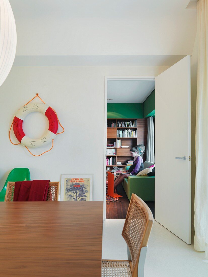 Chair with wicker mesh on the backrest in front of a wooden table in a dining room and a view through an open door of a lady sitting on a sofa