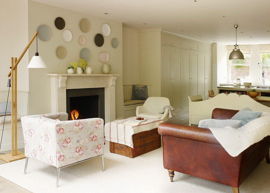 Armchair with floral upholstery and brown leather couch in front of open fireplace in open-plan, country-style interior