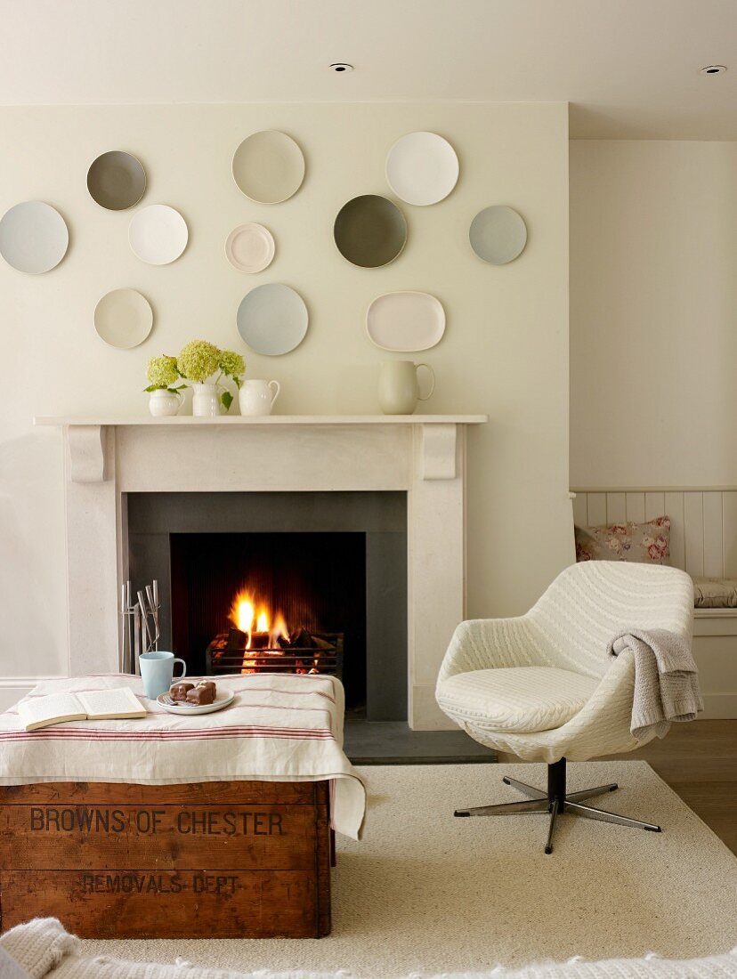 Vintage wooden trunk and pale swivel chair in front of open fire with decorative wall plates on chimney breast