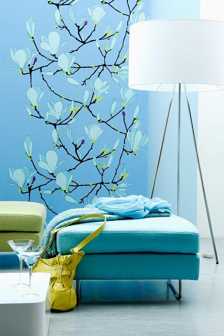 Ottomans in shades of aqua in front of designer standard lamp; blue wall-hanging with magnolia blossom pattern in background