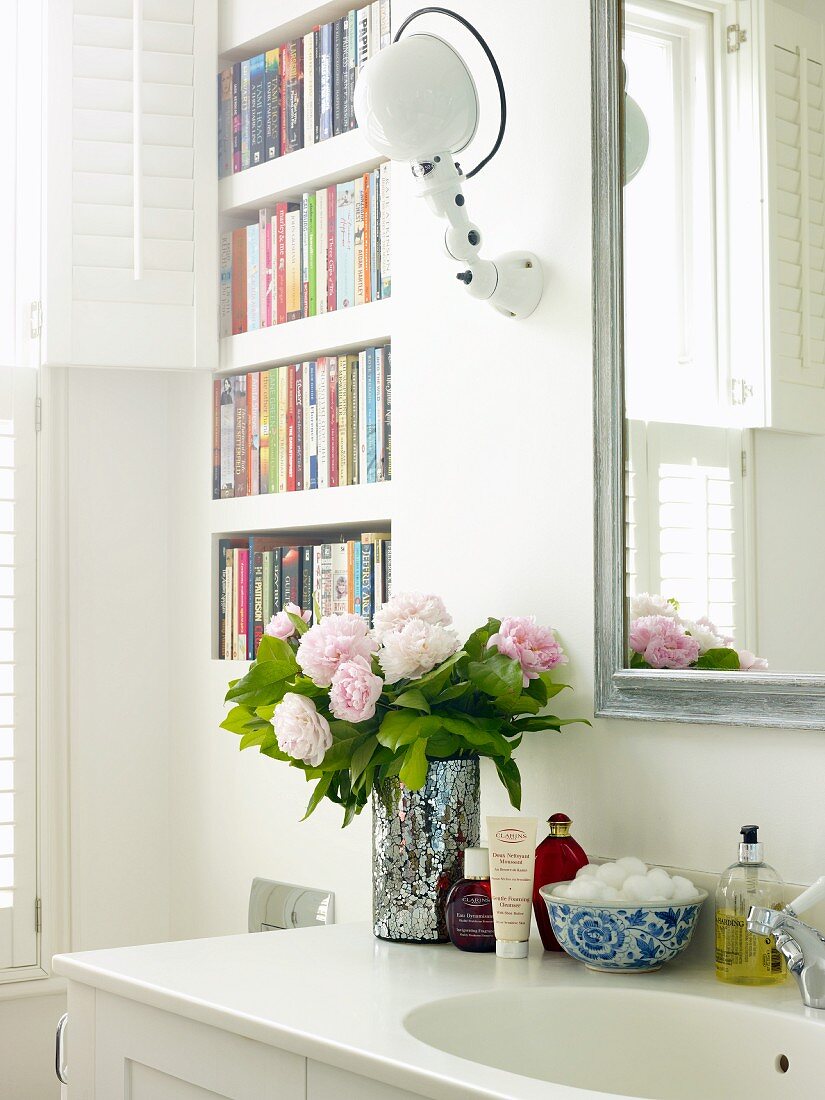Bouquet of peonies and bathroom utensils on washstand next to bookcase built into wall