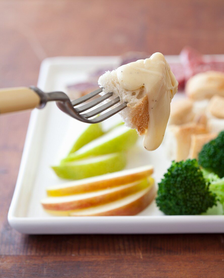 Gabel mit Brotstück und Käsefondue, Obst und Gemüse im Hintergrund