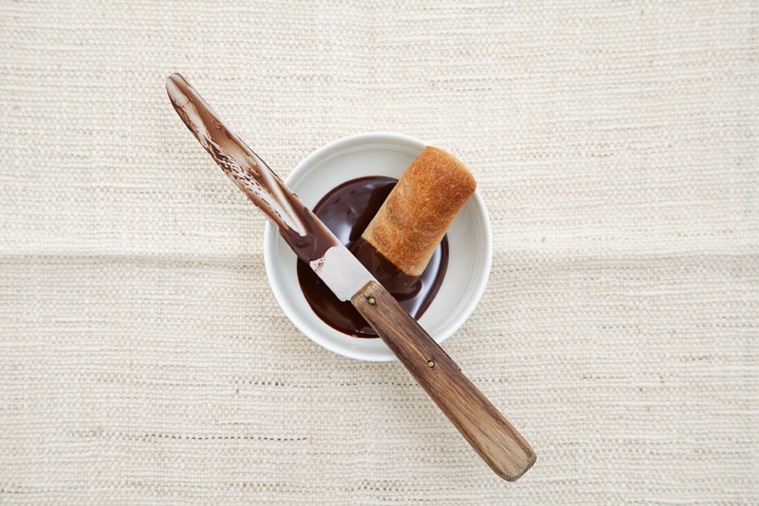 A bowl of melted chocolate with a piece of bread and a knife (seen from above)