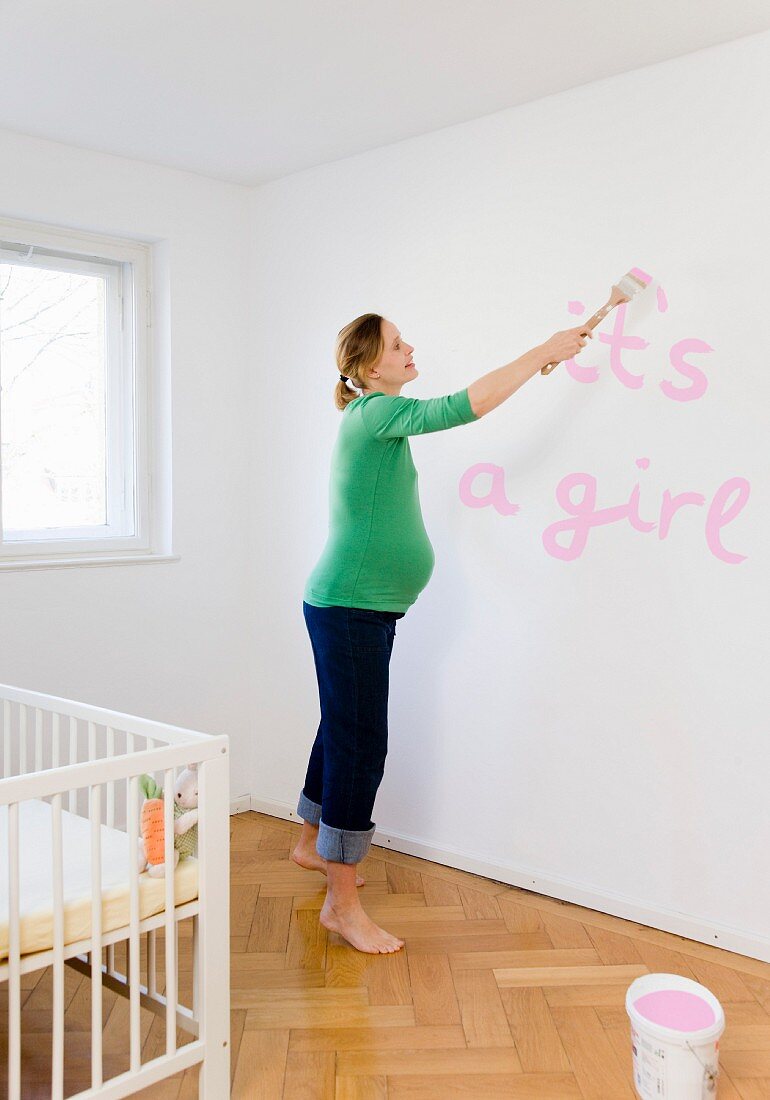 Schwangere Frau bemalt die Wand im Kinderzimmer
