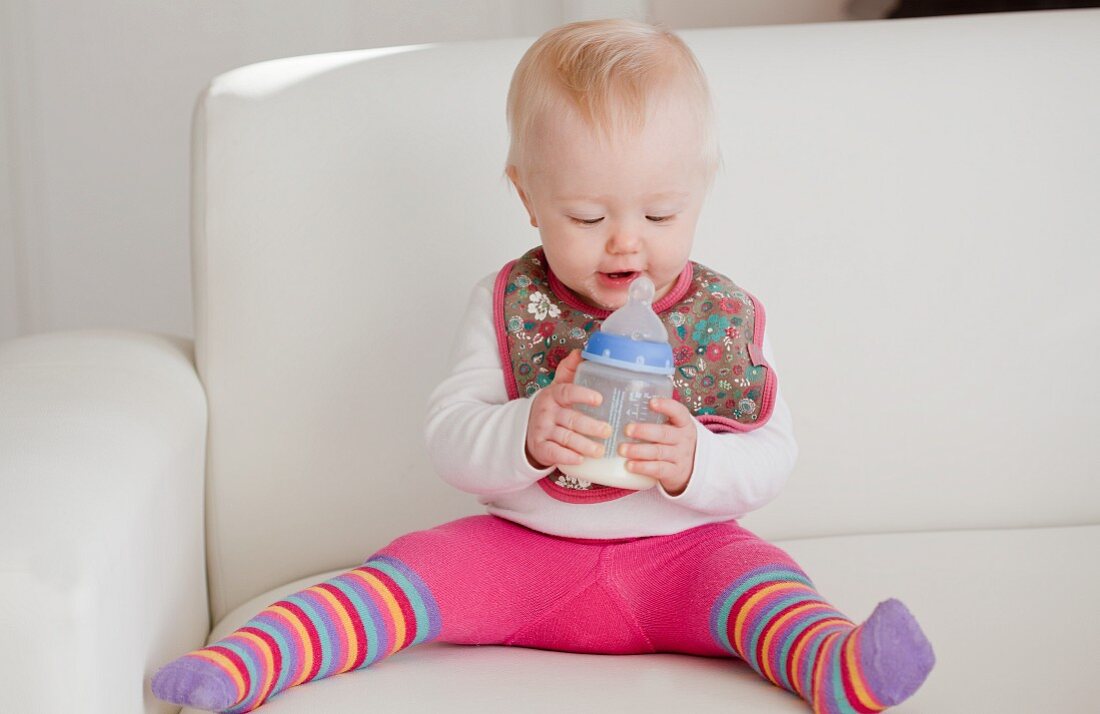 A baby sitting on a sofa with a bottle of milk