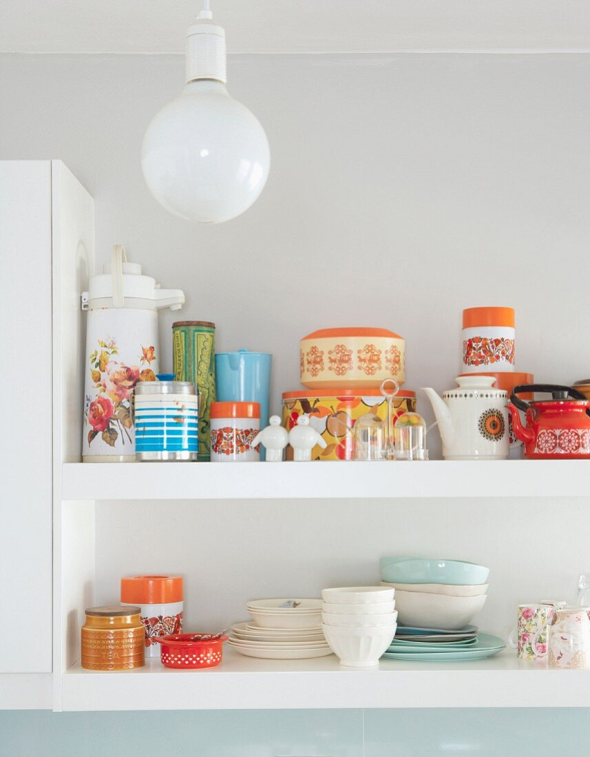 Detail of crockery and storage jars on white wall-mounted shelves behind spherical ceiling lamp