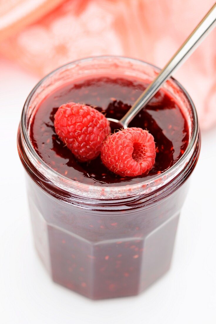 A jar of raspberry jam with a spoon and fresh raspberries