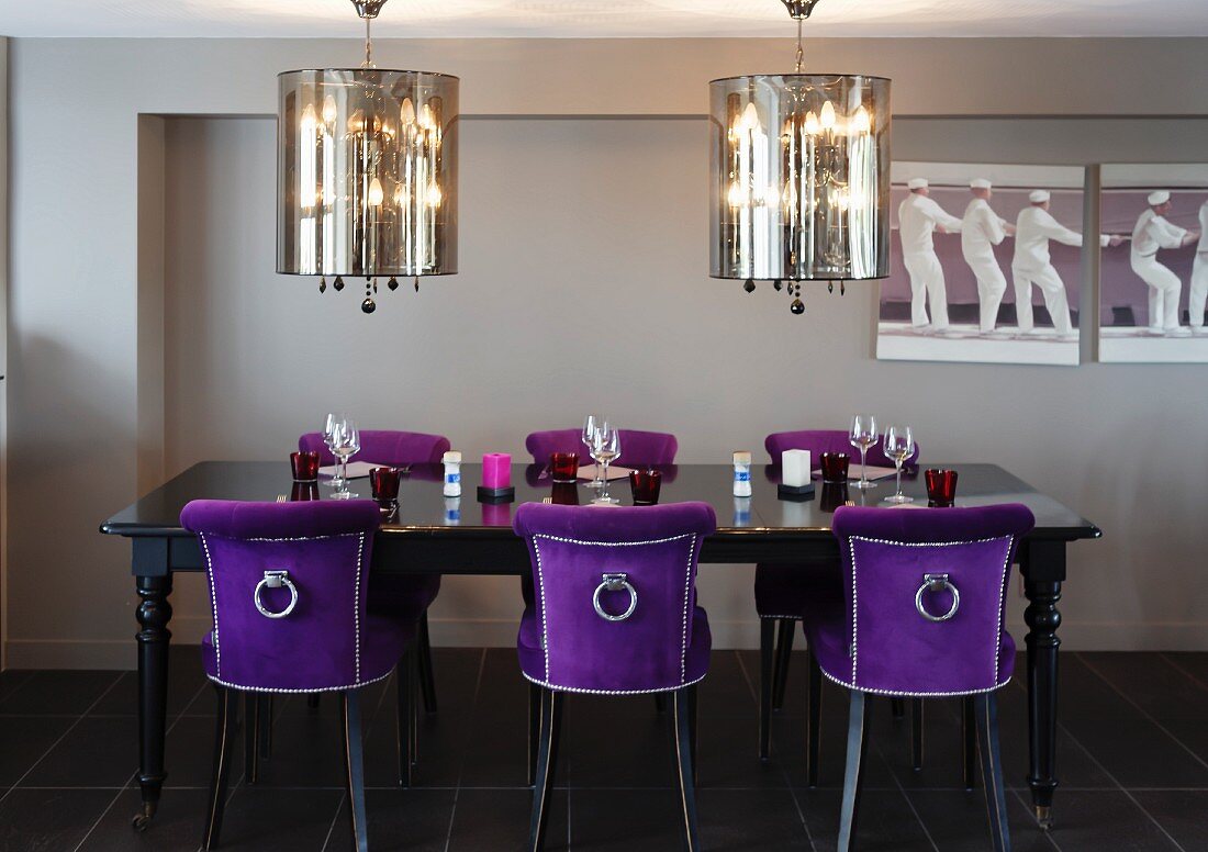 Opulent dining area with silver plated hanging lights and chairs covered in violet velvet at a set, black antique table