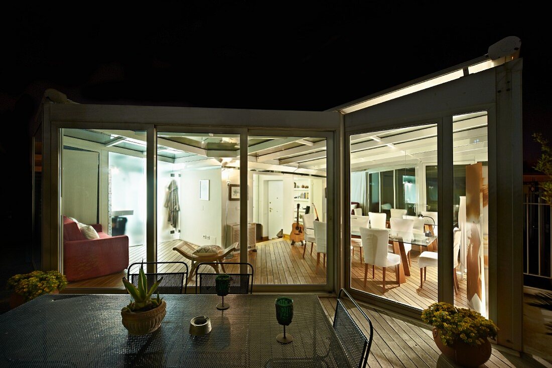 Night-time view from terrace with garden table into illuminated living area of apartment