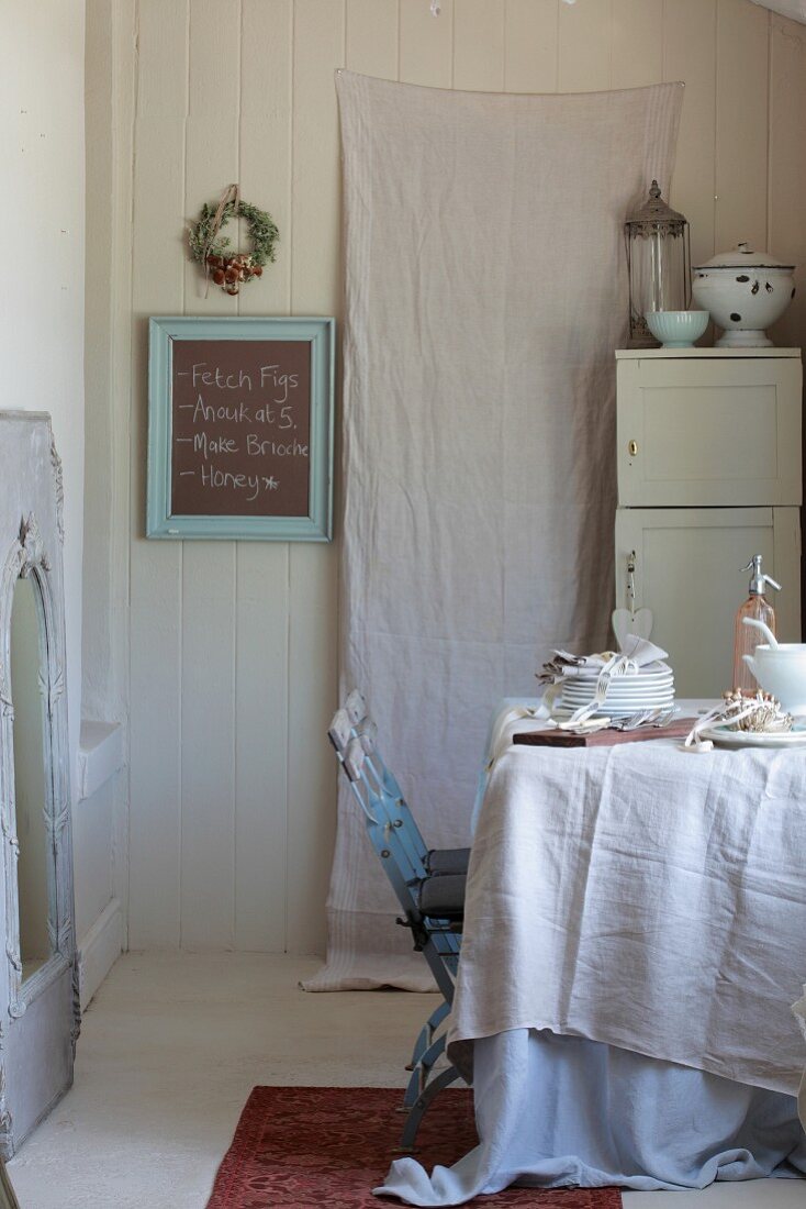 Linen cloths and crockery on table in vintage kitchen with white-painted cupboard and blackboard