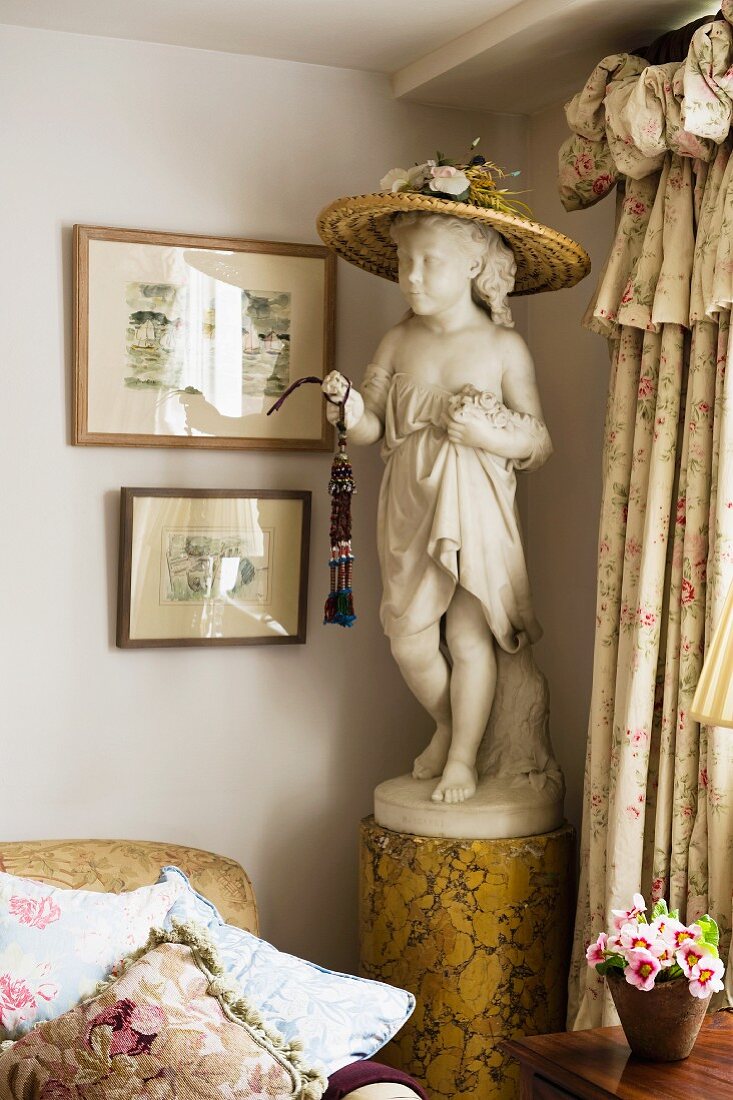 Mädchenstatue mit Strohhut auf Sockel in einer englischen Wohnzimmerecke neben Volantvorhang und Polstersofa mit vielen Kissen