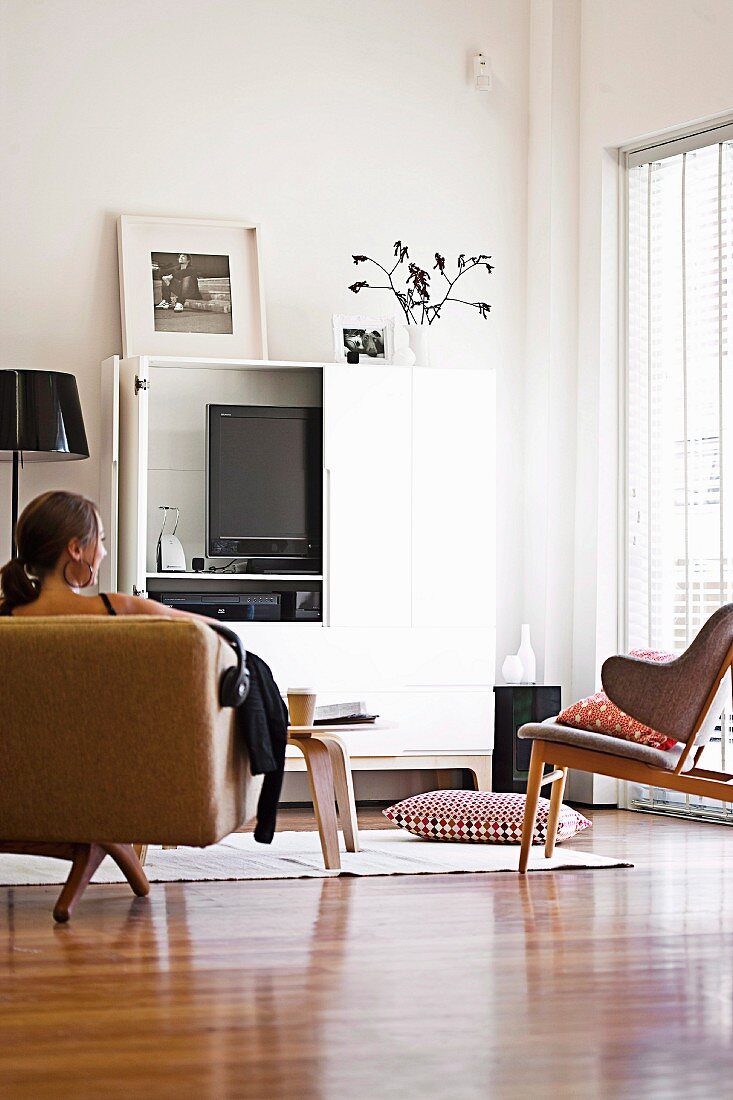 Wohnzimmerecke mit weiß lackiertem Schrankelement und Bildschirm, davor sitzende Dame auf Sitzmöbel im Fiftiesstil