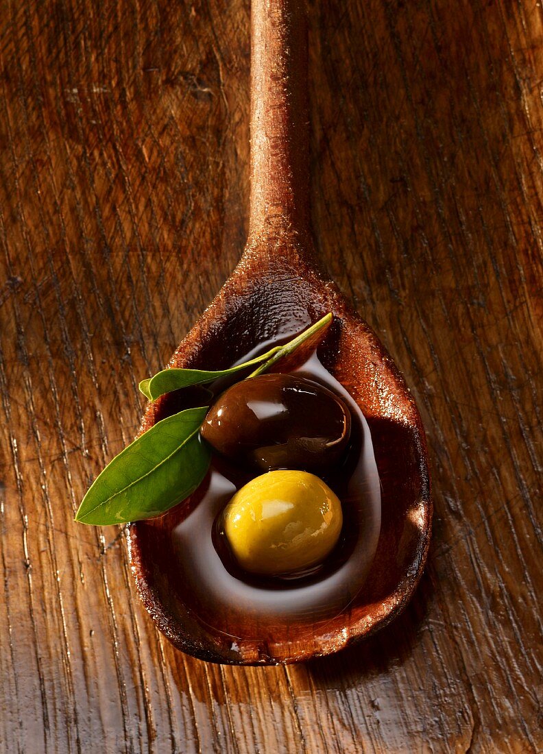 A black and green olive and an olive leaf in olive oil on a wooden spoon