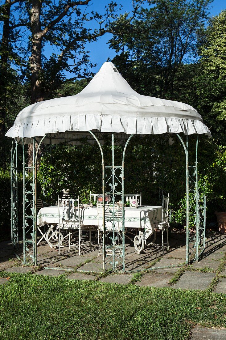 Climber-covered garden pavilion with white roof above set dining table and chairs in sunlight