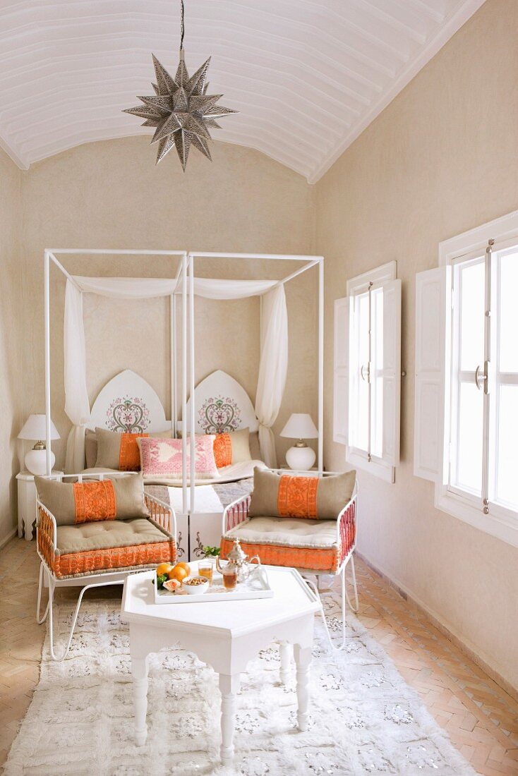 Bright, Oriental bedroom with white side table and set tea tray; ornate metal chairs with orange and beige cushions for taking afternoon tea