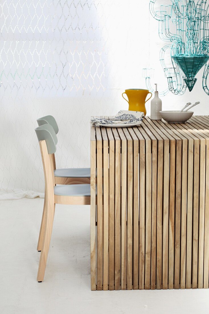 Wooden table made from many untreated wooden slats and grey chairs in front of white curtain