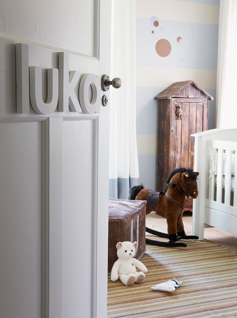 View into nursery with nostalgic atmosphere; white cot, rocking horse and small cupboard with roof against wall painted with pale blue and white stripes and circles