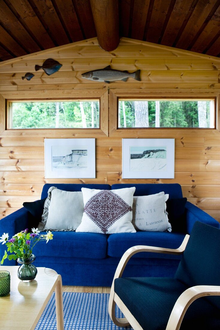 Gemütliche, elektrischblaue Sitzgruppe in schlichtem Holzhaus; über der Couch zwei Fensterschlitze und Fischtrophäen