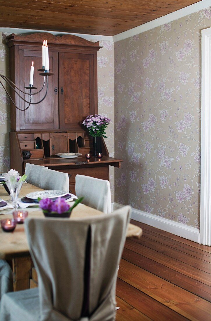 Antique bureau used as dining room dresser in room with subtle floral wallpaper; rustic dining table and chairs with loose linen covers in foreground