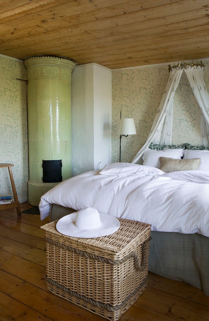 Lavish bed with canopy in pleasant room with many wooden elements, green patterned wallpaper and round tiled stove in corner