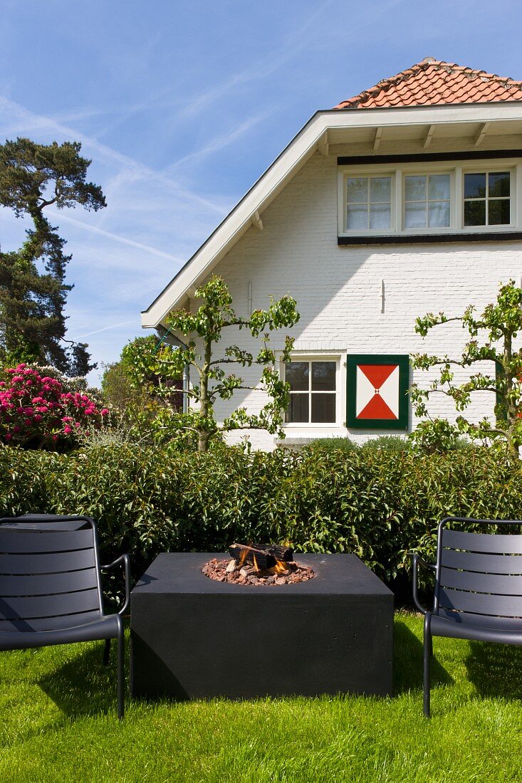 Minimalist table with fire pit and black garden chairs on the grass in front of a traditional country home