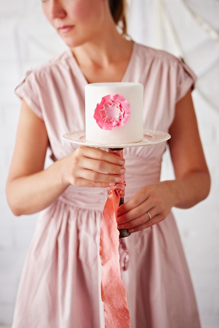 Woman Holding a Simple White Cake with Pink Flower Decoration on a Cake Plate