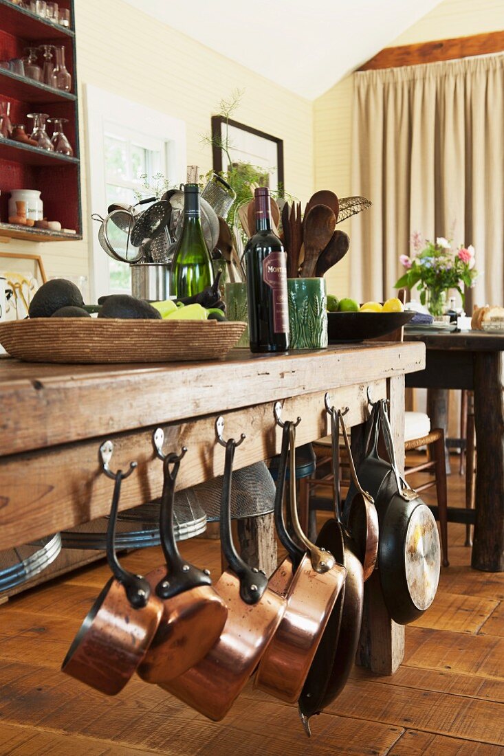 Massive wooden table with copper pans hanging from one side and set with shallow basket, wine bottles and storage containers in dining area on rustic wooden floor