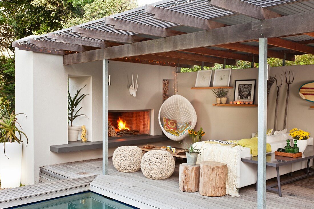 Seating area on roofed terrace next to pool with open fireplace, hanging chair & stools made from natural materials