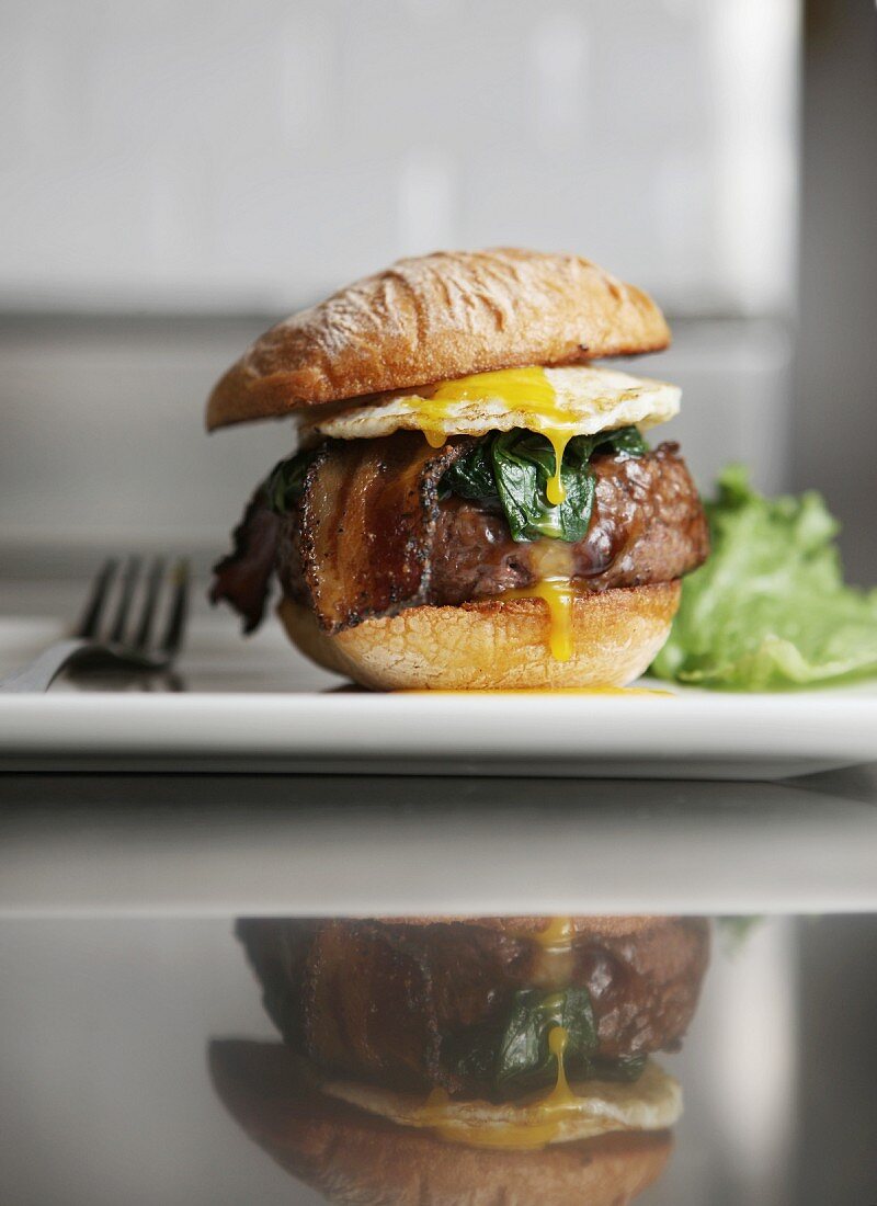 A Hamburger with Fried Egg, Bacon and Spinach on a White Plate