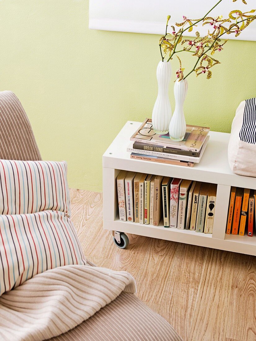 Blumenvasen auf Bücherregal mit Rollen vor pastellgrüner Wand und Kissen auf Sessel