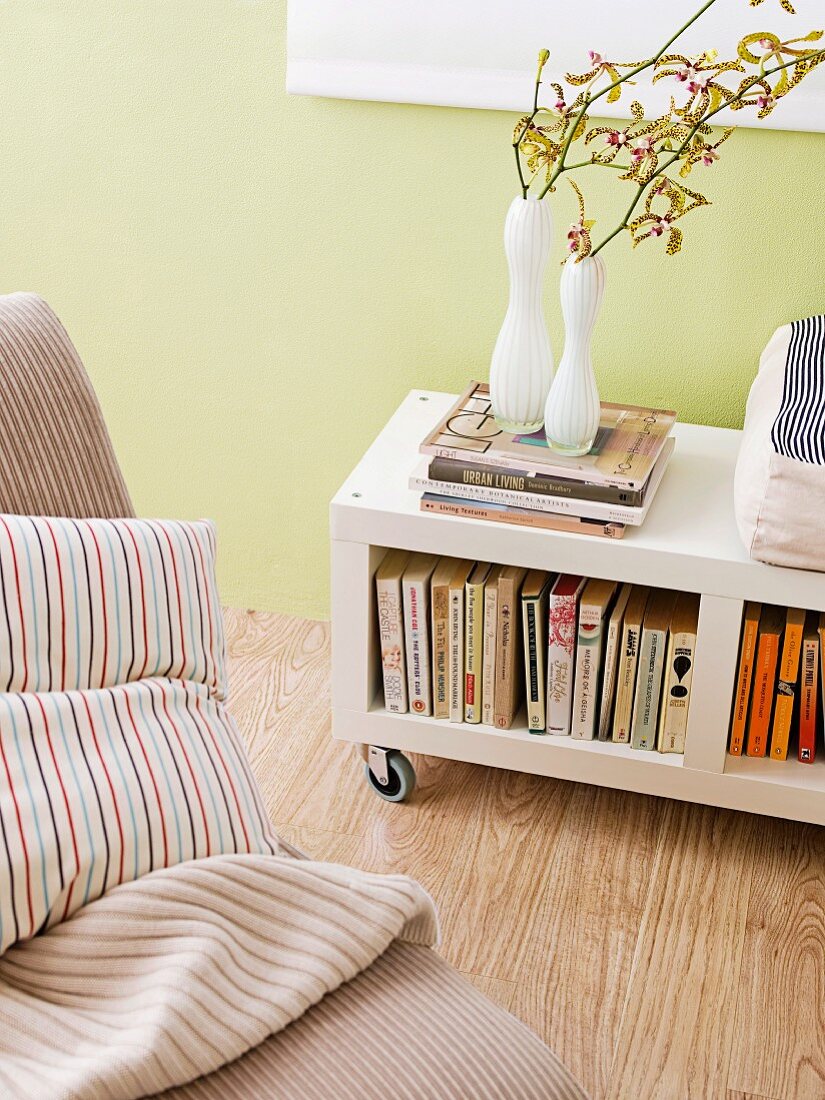 Vases of flowers on bookcase with castors against pastel green wall and scatter cushions on armchair