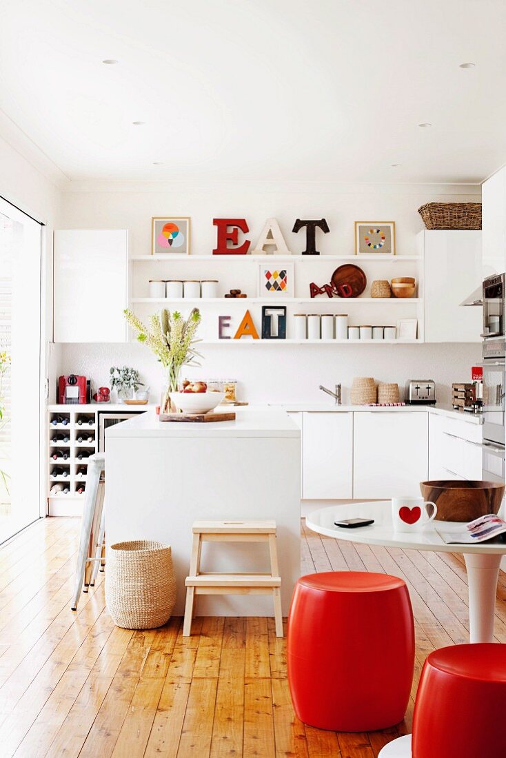 White kitchen with kitchen island and wooden floor; red, barrel-shaped stools at round table in foreground, fitted cupboards and ornaments on wall-mounted shelves in background