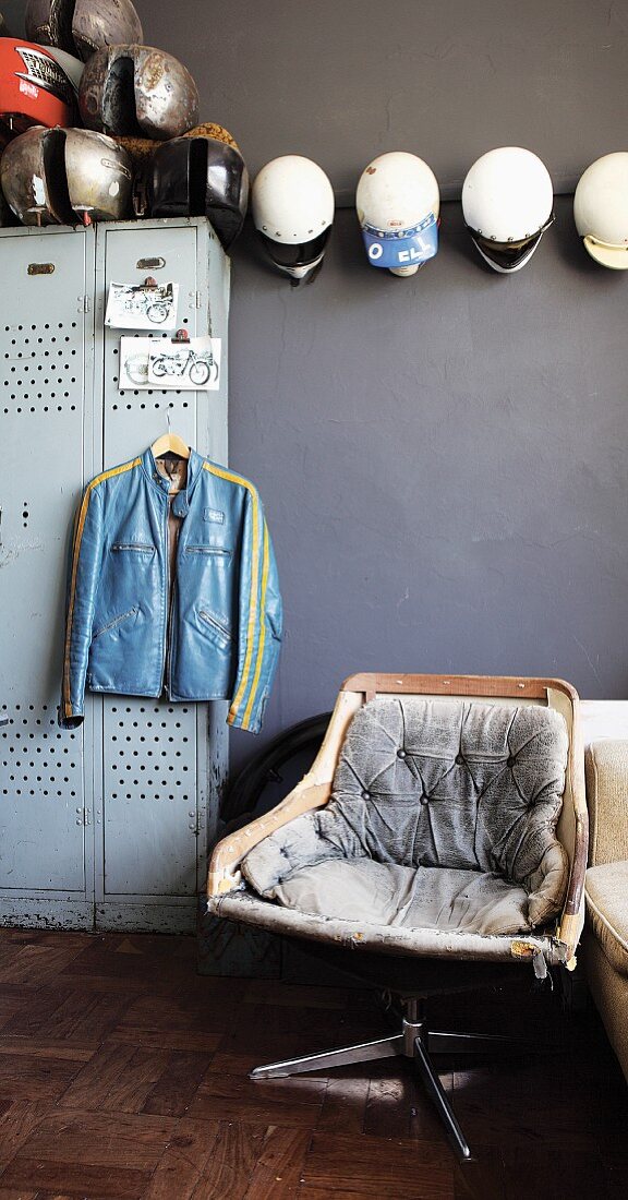 Vintage swivel chair in front of motorbike jacket hanging on locker and collection of helmets on grey wall