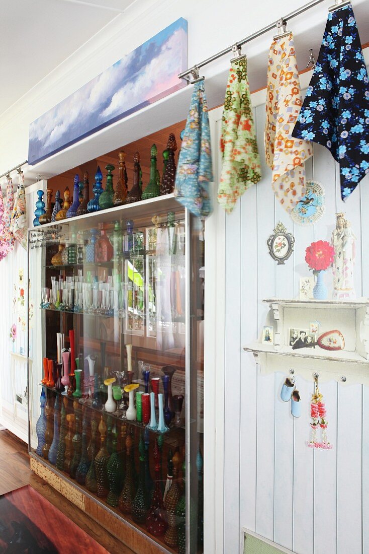 Collection of coloured glass bottles in partition shelving between patterned cloths hanging from clips and surrounded by kitsch