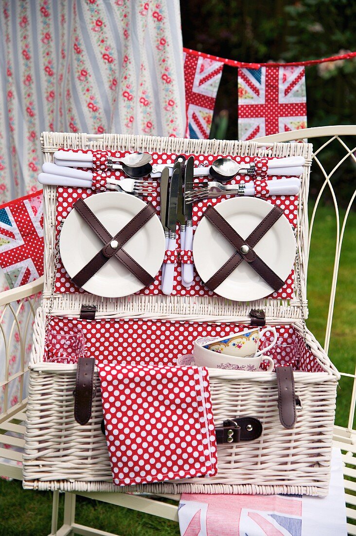 Plates and cutlery in the lid of an open, white picnic basket lined with red and white spotted fabric and stylised Union Jack bunting in the background