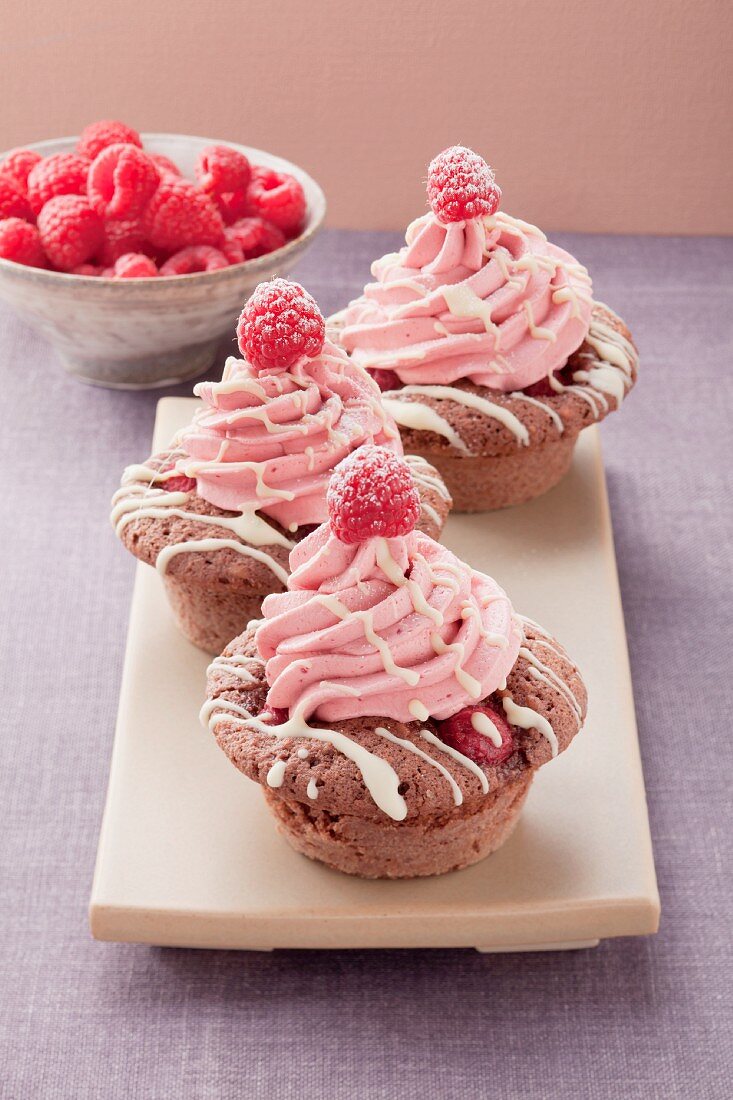 Drei Cupcakes mit Himbeercreme, Schale mit Himbeeren im Hintergrund