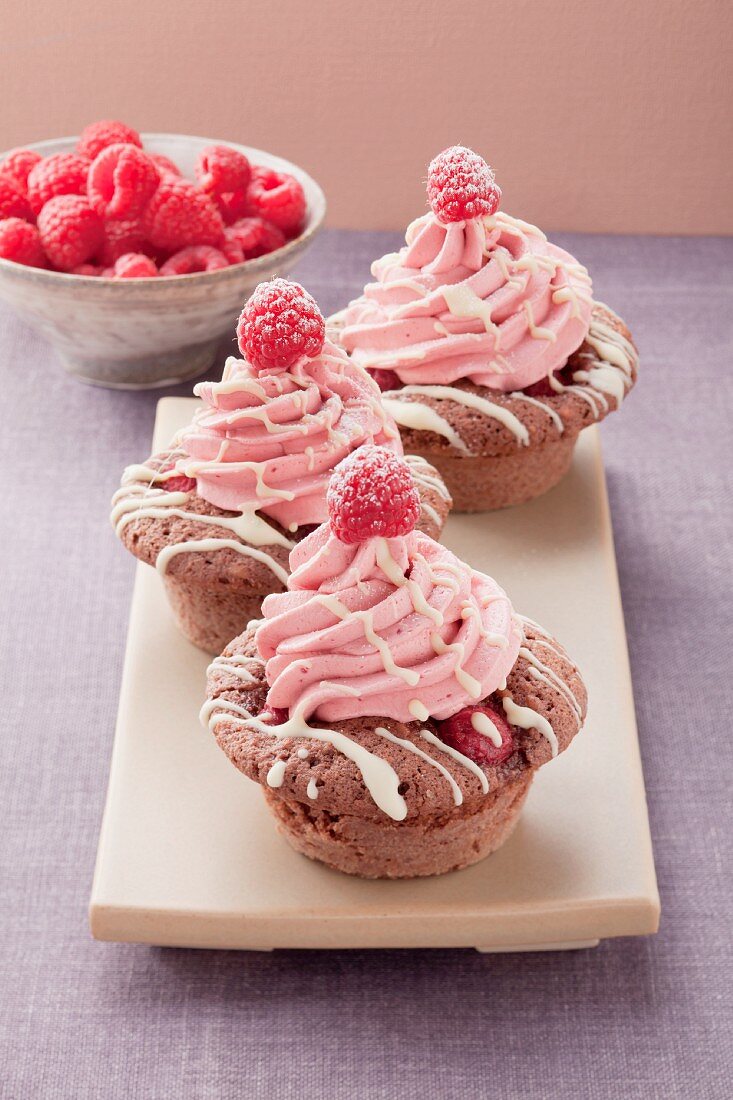 Three cupcakes with raspberry cream topping, with a bowl of raspberries in the background
