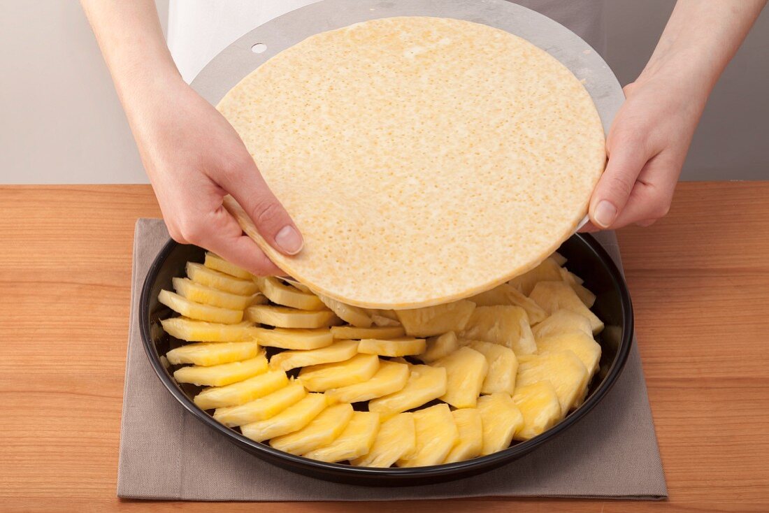 Hands laying the cake base onto pineapples in a baking tin