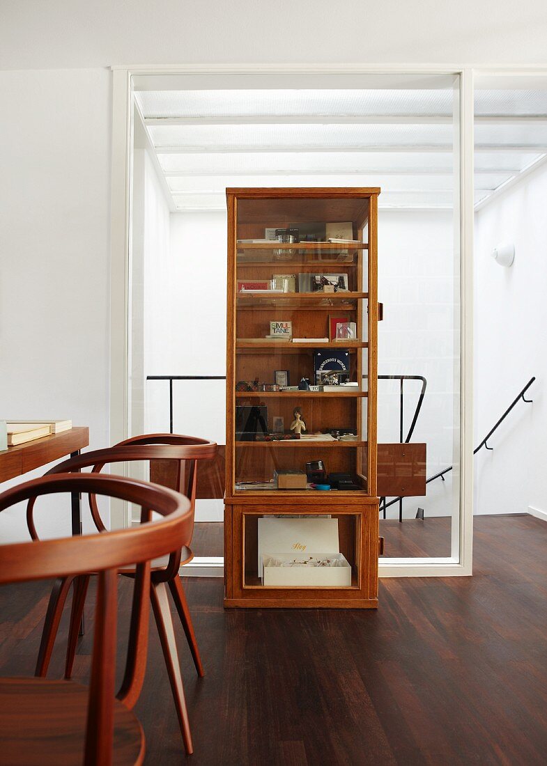 Wooden display case against glass wall element leading to landing on dark, wooden parquet flooring with table and elegant, dark wooden chairs in foreground