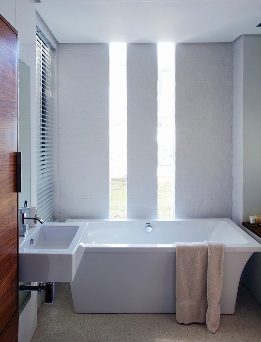 Elegant, minimalist bathroom with exotic wood door and vertical window strips behind free-standing, designer bathtub