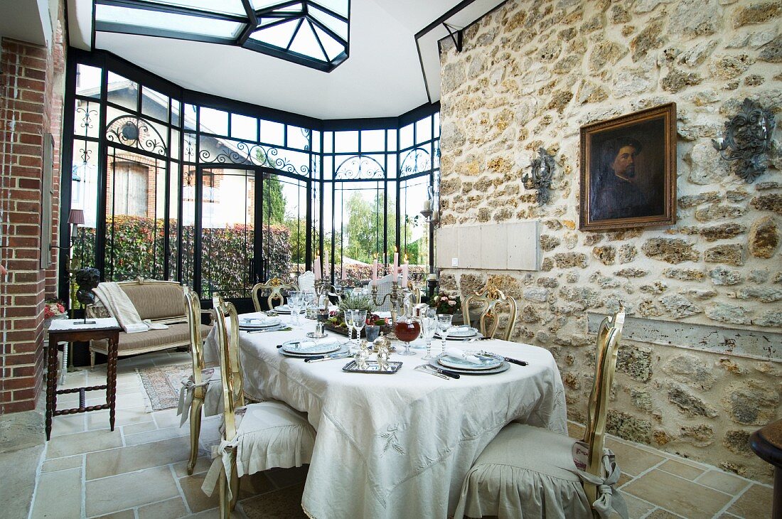 Rustic wall in bright dining room with huge, glass bay window; long, set dining table and gilt, Baroque chairs with romantic loose covers in foreground