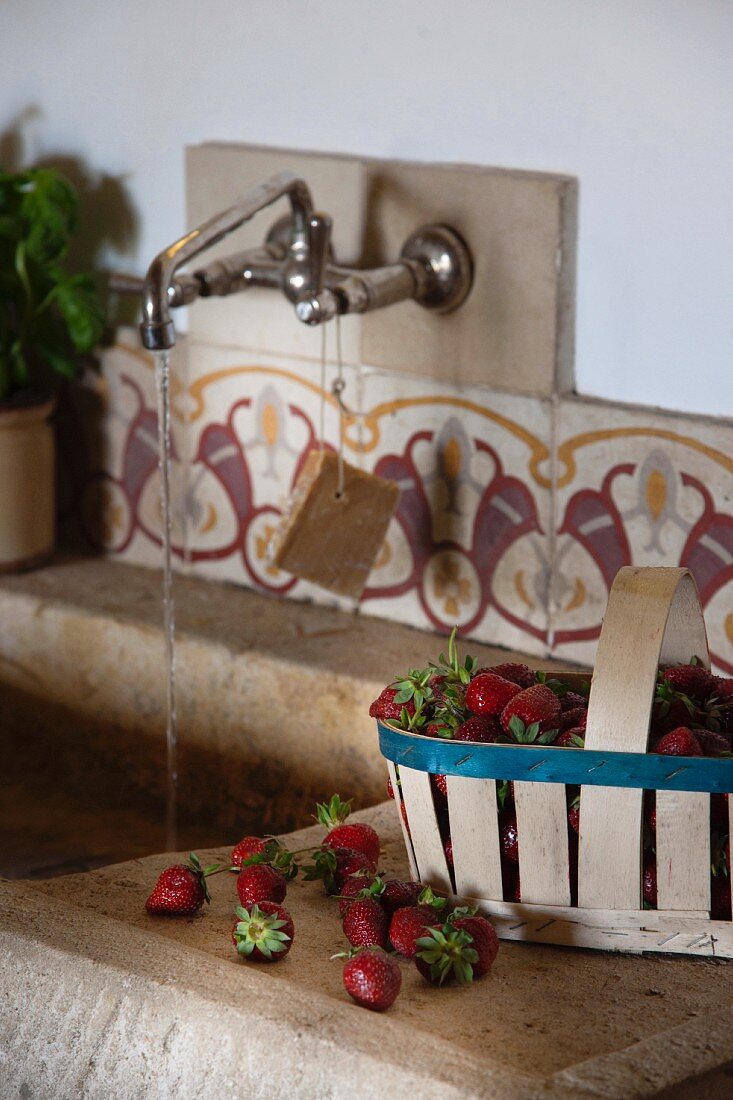 Chip wood basket of strawberries on side of stone sink; water running from wall-mounted taps above old, patterned wall tiles