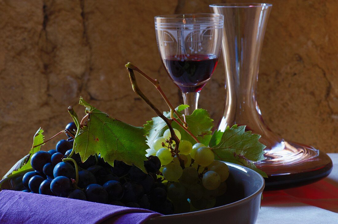 A bowl of red and green grapes in front of a decanter and a glass of red wine