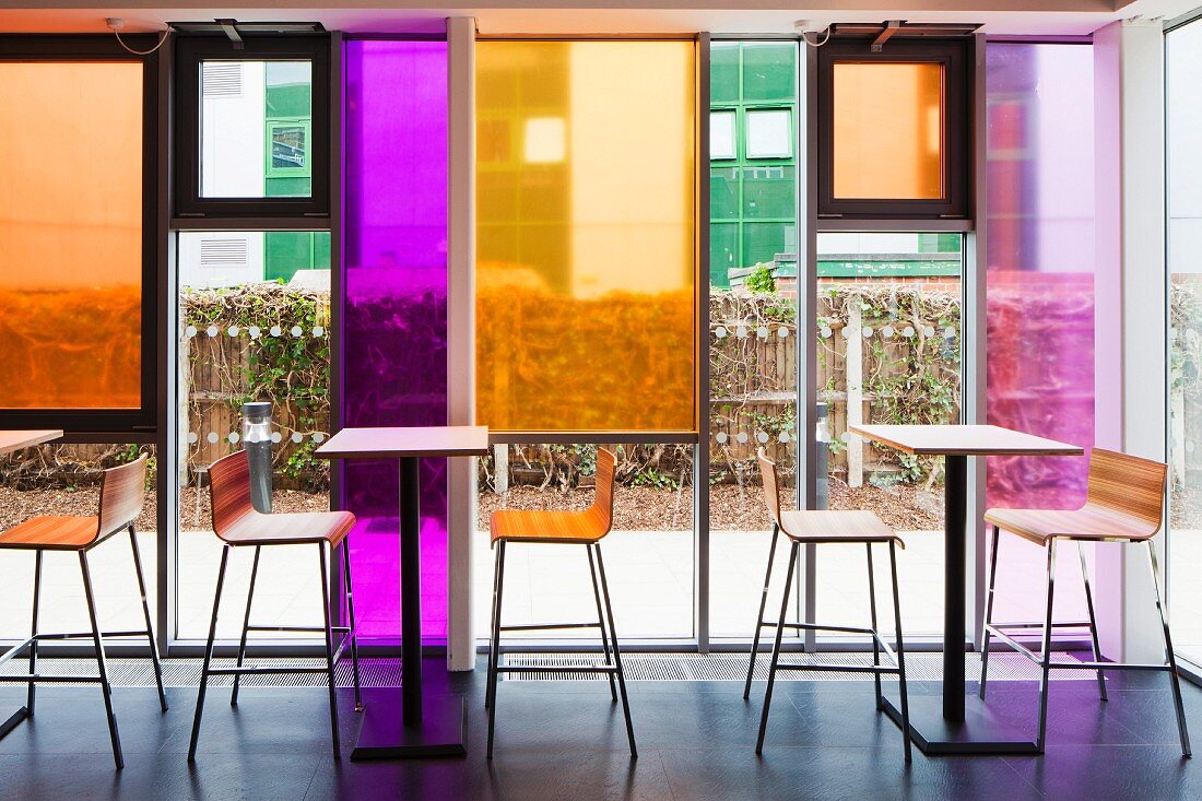 Tall tables and bar stools in front of glass facade with coloured glass panels