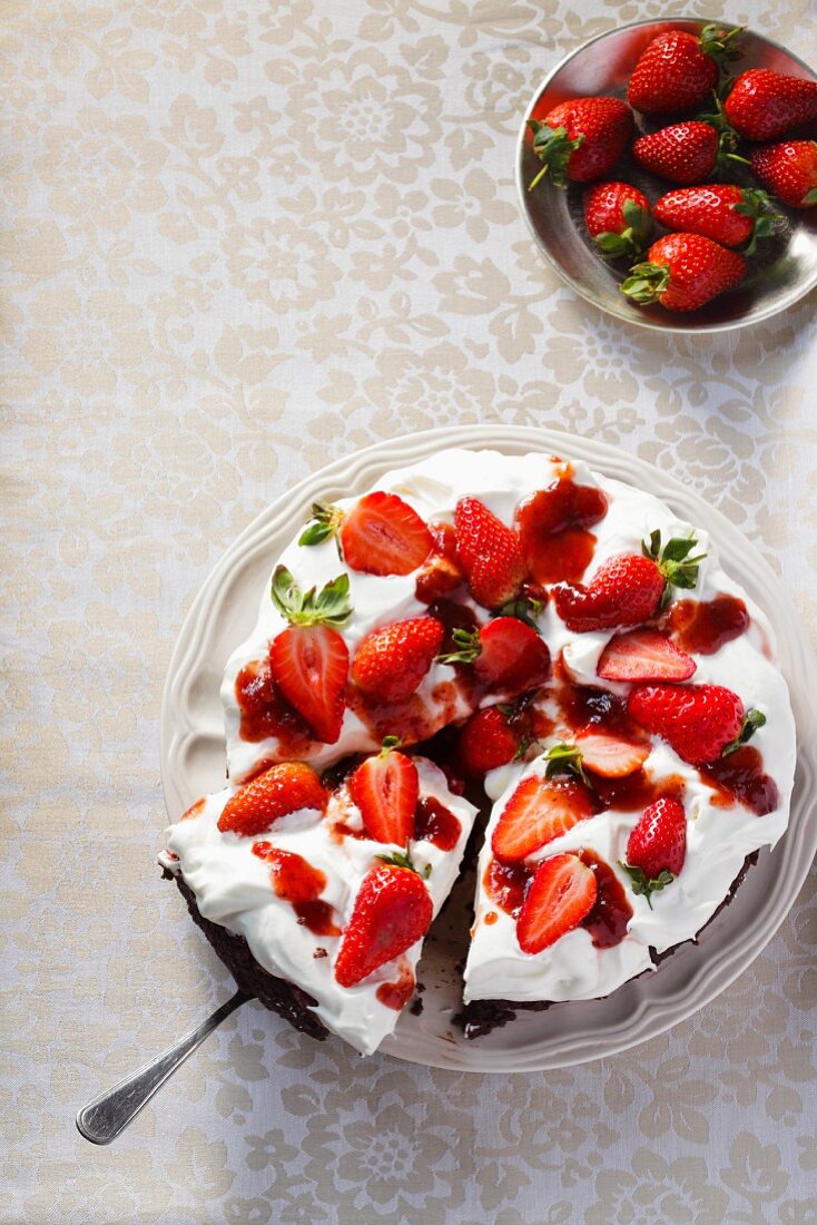 A cream cake topped with strawberries with a bowl of fresh strawberries next to it; seen from above