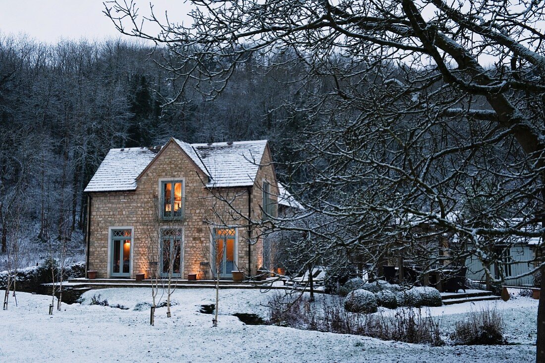 View across snowy stream of caretaker's house belonging to English stately home; twilight scene with lights in windows
