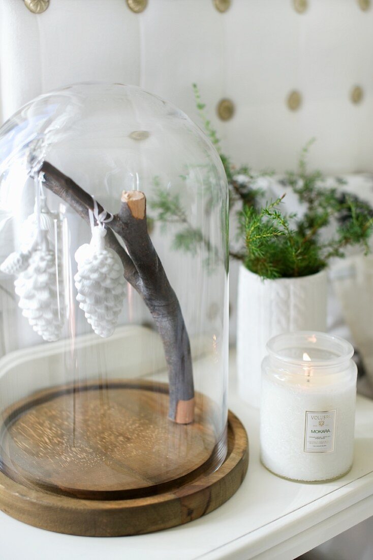 Stylised fir cones hanging from branch under glass cover and lit candle lantern on bedside table
