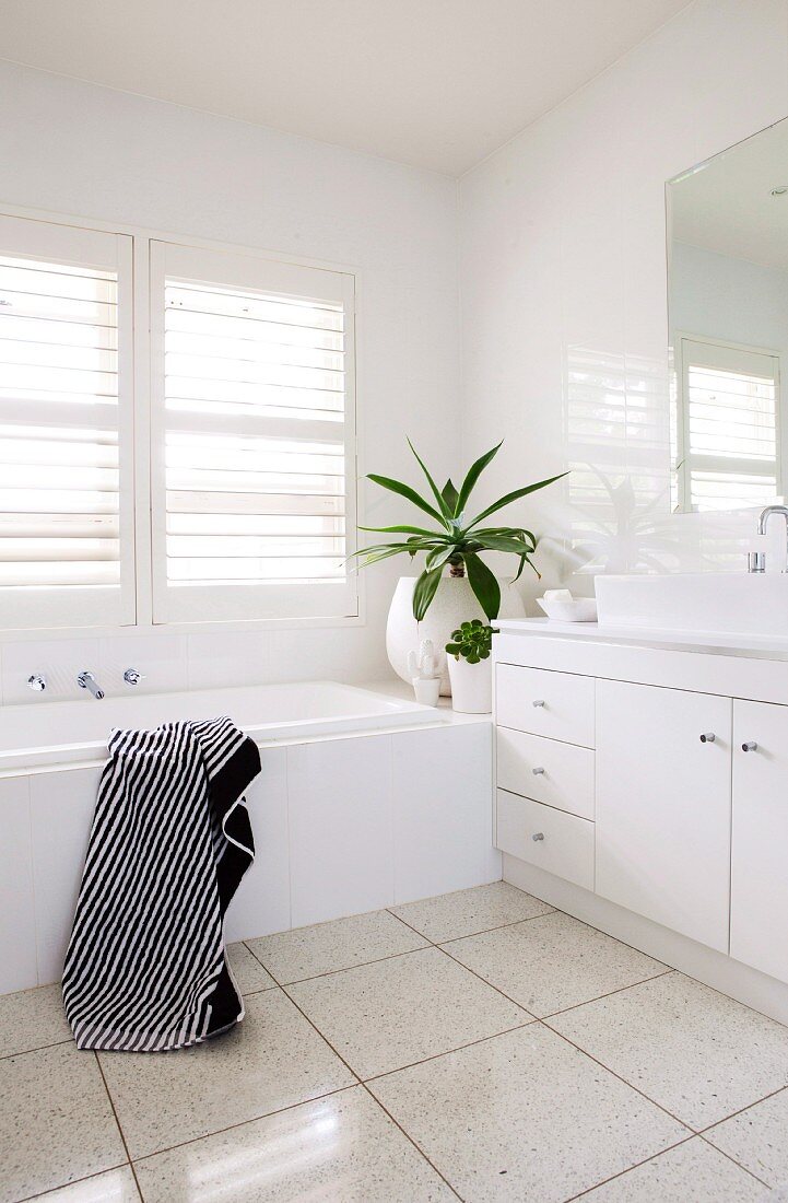 Weisser Waschtisch und Badewanne in Badezimmerecke vor Fenster mit Innenläden aus weissen Holzlamellen